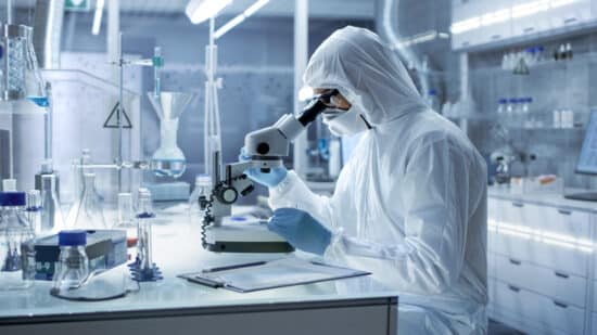 In a Secure High Level Research Laboratory Scientist in a Coverall Examines Petri Dish Under Microscope.