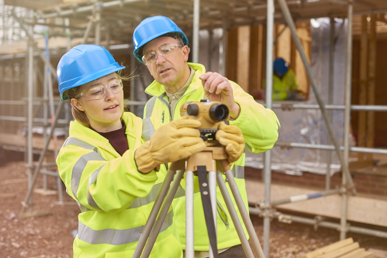 a female construction worker stands behind a builder's level on a building site .Behind her a co-worker walks across the development