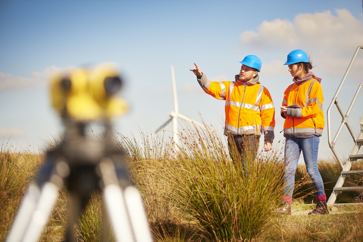 two wind farm engineers using a builder's level to plan out the expansion of the wind farm site. they are wearing orange hi vis jackets and blue hard hats . one is male , one is female. In the foreground the female is looking through the level whilst the male engineer is approaching .In the background wind turbines can be seen across the landscape.