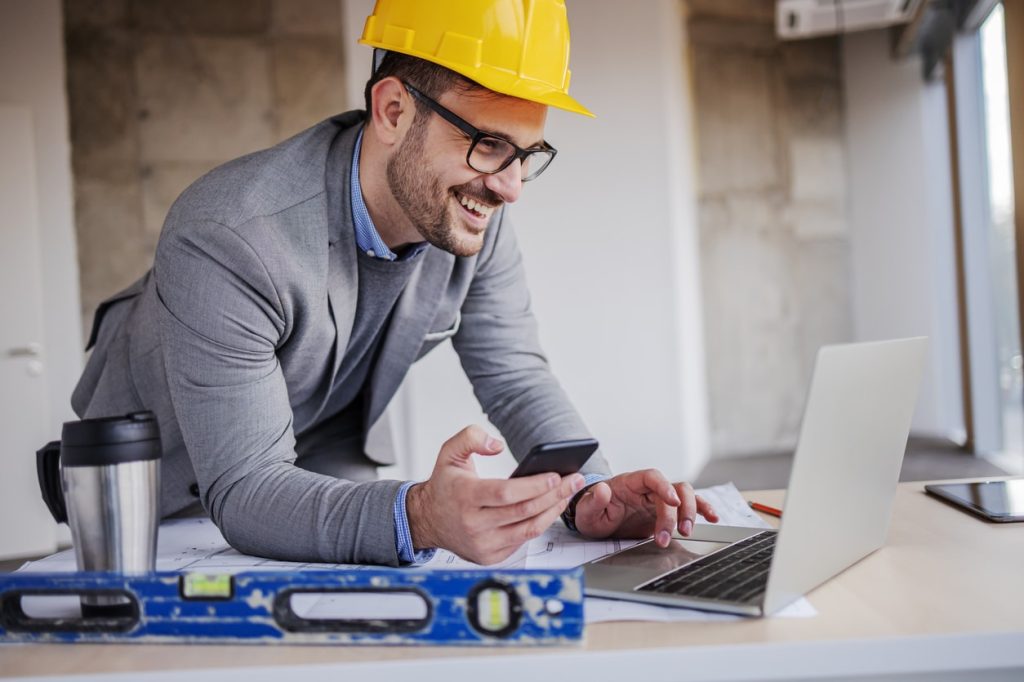 Young smiling architect in suit with helmet on head holding smart phone and using laptop while leaning on table in building in construction process.