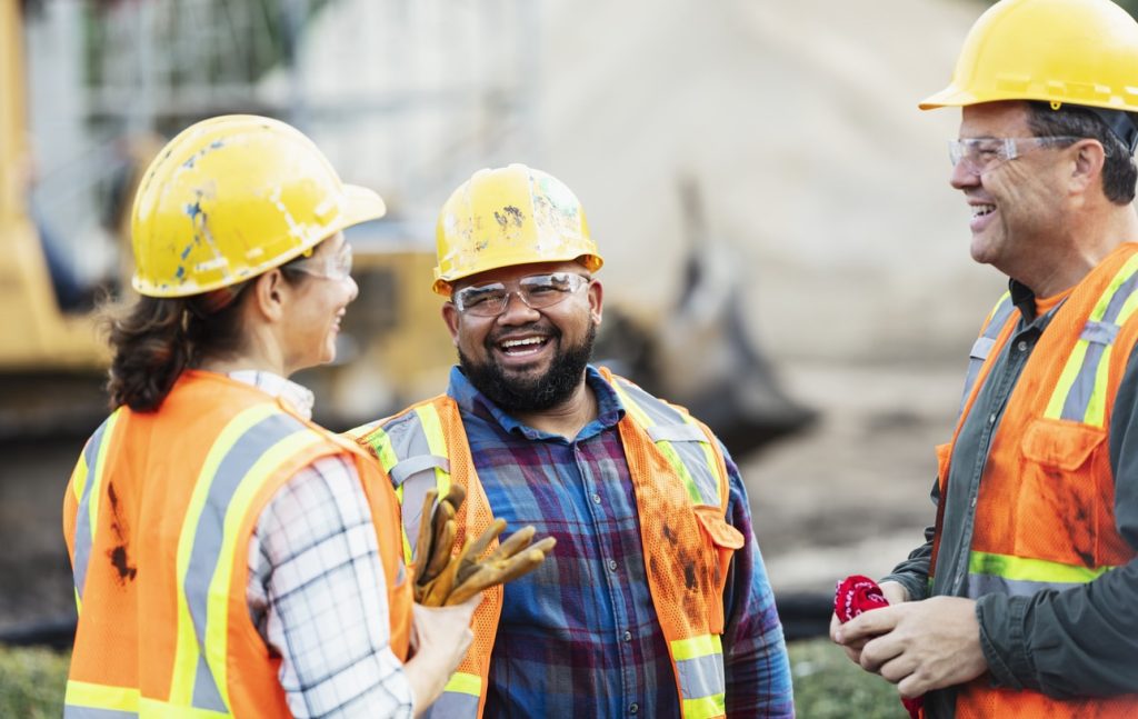 A group of three multi-ethnic workers at a construction site wearing hard hats, safety glasses and reflective clothing, smiling and conversing.